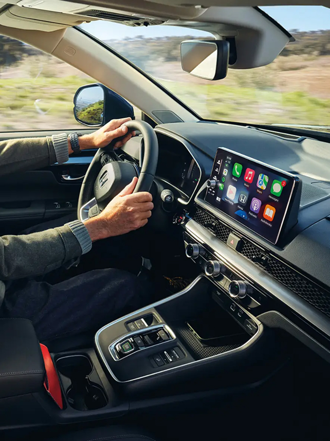 Vue intérieure d'une voiture moderne avec un conducteur assis au volant, montrant le tableau de bord, le volant, la console centrale et un large écran tactile affichant des icônes d'applications colorées, tandis qu'un paysage de campagne défile par les fenêtres.