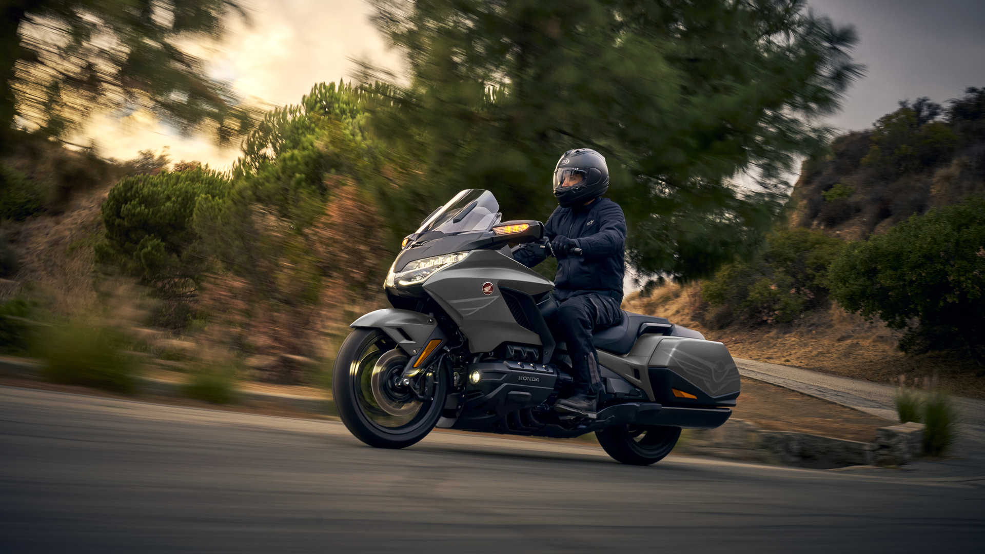 A Honda Gold Wing motorcycle being ridden on a curving road at dusk. The large touring bike is gray in color, with a rider wearing dark clothing. Trees and foliage line the road, creating a scenic backdrop.