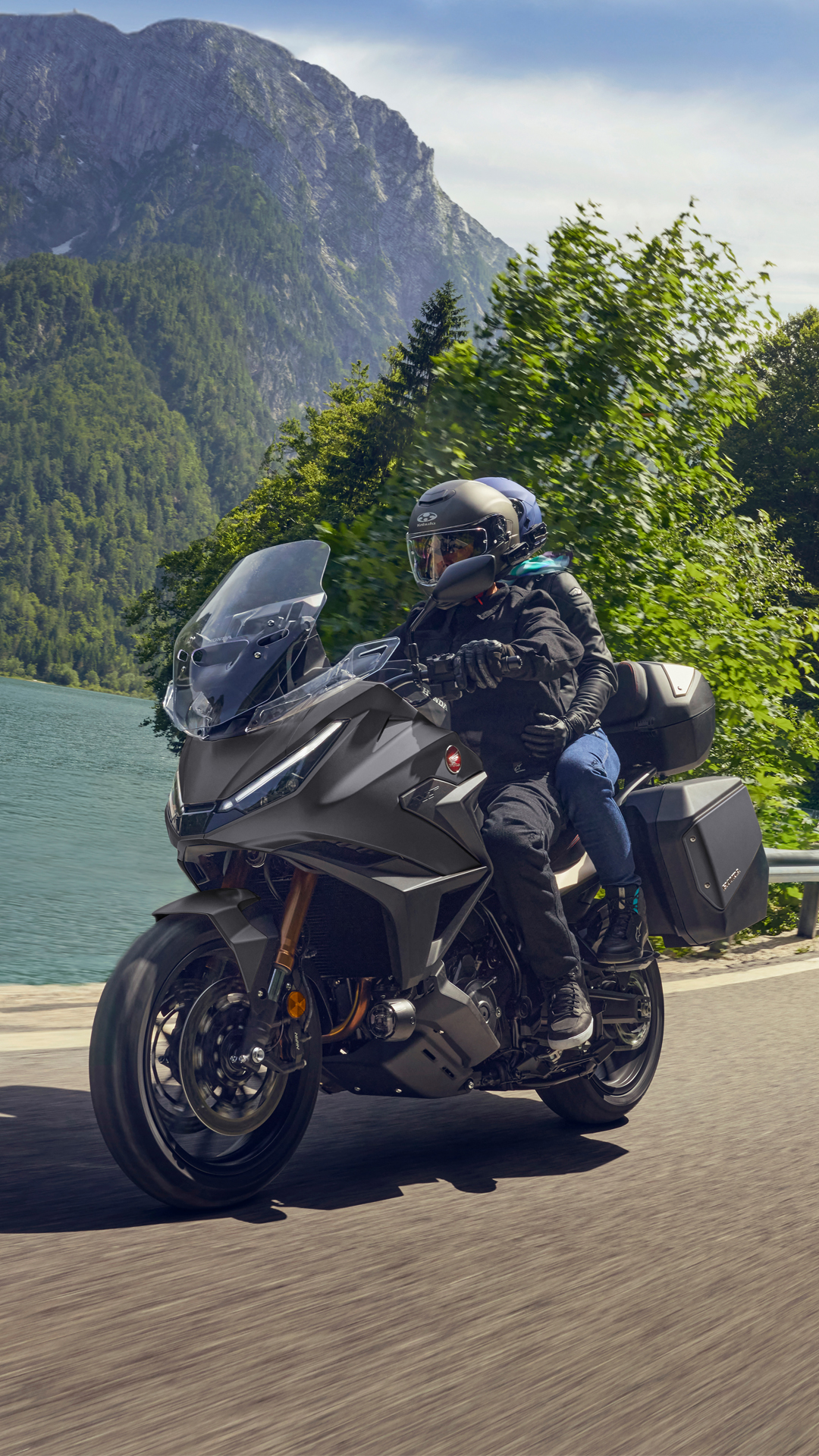 A Honda NT1100 touring motorcycle rides along a scenic mountain road. The bike is gray and equipped with side cases. In the background, a steep mountain face rises above a turquoise lake, with lush green trees in the foreground.