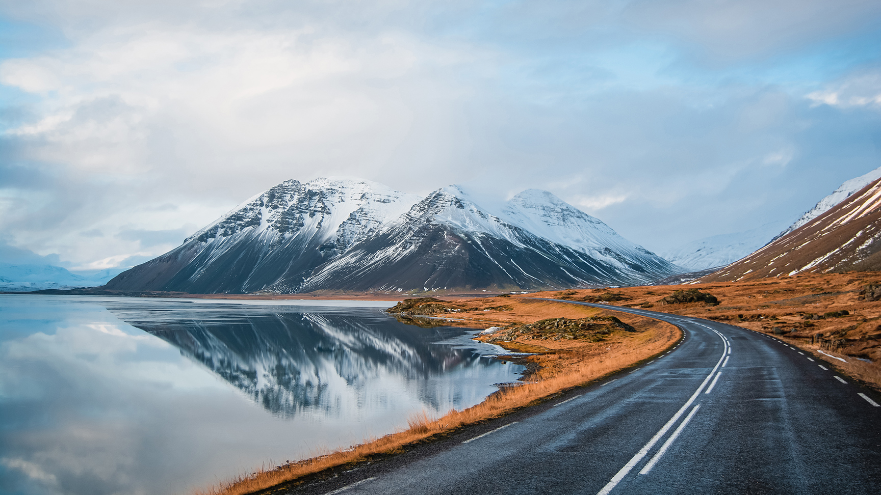 Photo hivernale panoramique d’une route jonchant la côte du lac jusqu’à des montagnes volcaniques. De hauts pics rocheux vêtus d’un manteau de neige dont le reflet est visible à la surface de l’eau. Point de vue d’un conducteur sur la rocade, Islande.