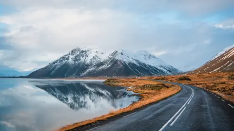 Photo hivernale panoramique d’une route jonchant la côte du lac jusqu’à des montagnes volcaniques. De hauts pics rocheux vêtus d’un manteau de neige dont le reflet est visible à la surface de l’eau. Point de vue d’un conducteur sur la rocade, Islande.