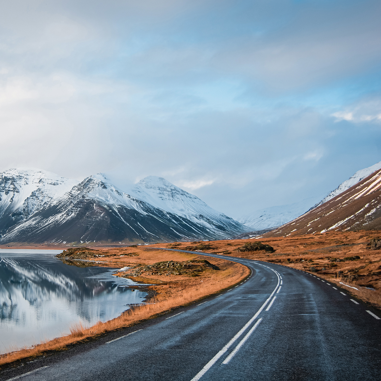 Photo hivernale panoramique d’une route jonchant la côte du lac jusqu’à des montagnes volcaniques. De hauts pics rocheux vêtus d’un manteau de neige dont le reflet est visible à la surface de l’eau. Point de vue d’un conducteur sur la rocade, Islande.