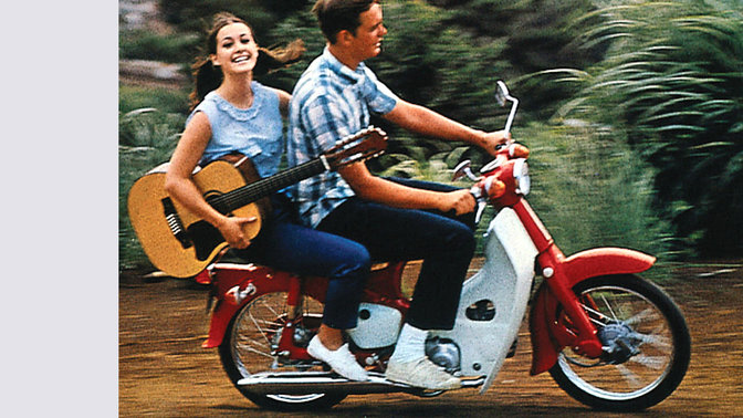 Honda Cub avec pilote et passager portant une guitare. Sur chemin de terre. Vue de face droite.